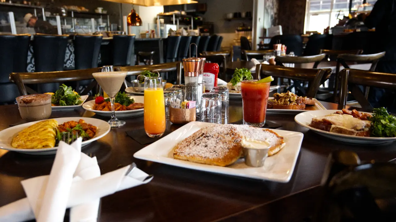 A full restaurant table featuring an array of artfully plated brunch dishes and fresh cocktails, set against a warm, stylish bistro dining room.