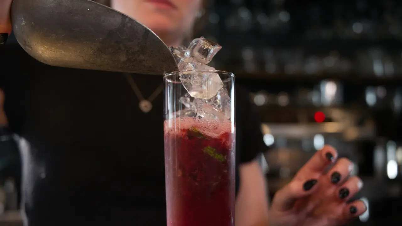 Close-up of a mixologist pouring fresh ice into a highball glass containing a handcrafted berry and mint cocktail at an upscale bar.