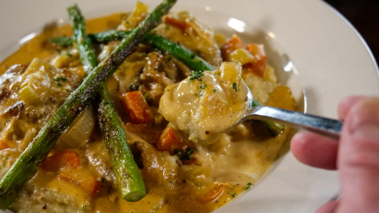 Close-up of a diner lifting a spoon full of creamy sauce from an artfully presented entrée with grilled asparagus spears and root vegetables.